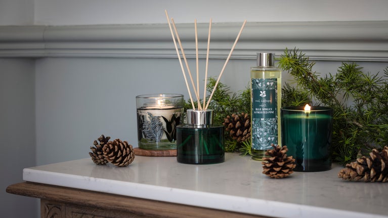 Autumn home fragrance collection with candles, reed diffuser, pine cones and evergreen foliage on a marble-topped table.